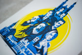 A close-up of a printed artwork featuring stylized blue portraits of historical figures and buildings arranged around a large yellow circle on a white background.