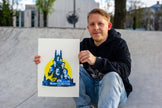 A man wearing a black hoodie and blue jeans sits on concrete at an outdoor skatepark, holding up a white art print featuring a blue graphic illustration of a castle and faces on a yellow background.