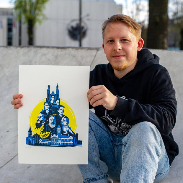 A man wearing a black hoodie and blue jeans sits on concrete at an outdoor skatepark, holding up a white art print featuring a blue graphic illustration of a castle and faces on a yellow background.